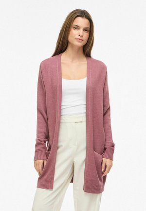 Moteris dėvi dulkinės rožinės spalvos atvirą kardiganą, baltą be rankovių palaidinę ir aukštakulnius baltus kelnes priešais paprastą foną.