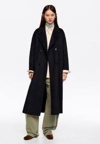 Mujer con un abrigo largo negro de doble botonadura, una camisa beige, un suéter gris de cuello alto, pantalones sueltos verde claro y zapatos marrones, de pie con una expresión neutral.