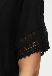 Μαύρο κοντομάνικο με φλοράλ δαντέλα που δείχνει το δέρμα από κάτω στο πάνω μέρος του βραχίονα ενός ατόμου, σε λευκό φόντο.