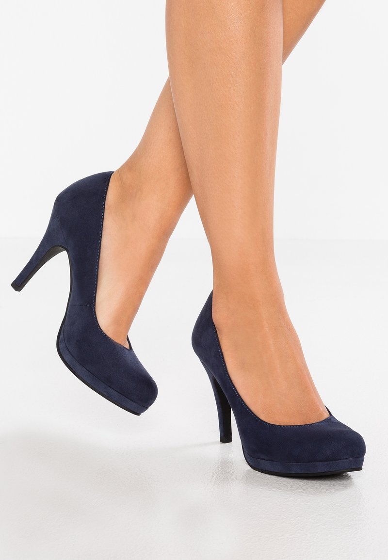 Jambes de femme portant des escarpins en daim bleu foncé à talons hauts, debout sur une surface blanche réfléchissante avec un pied légèrement levé.
