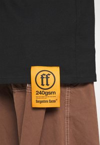 Sort bomulds T-shirt med et fremtrædende orange mærke, der viser "ff" logoet og teksten "240gsm", parret med brunt stof nedenfor.