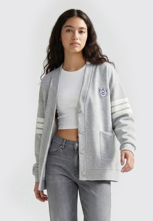 Ung kvinde med langt mørkt hår iført en lysegrå knap-op cardigan med hvide striber på ærmerne og en hvid cropped top, stående.