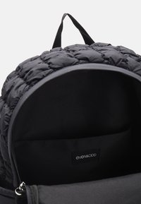 Mochila negra acolchada con parte superior redondeada, con un forro interior liso y una etiqueta de marca en el interior. Incluye un asa superior y cierre de cremallera.