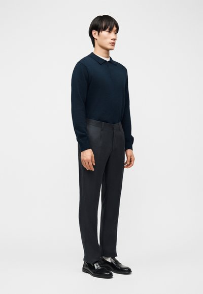 Άνδρας που στέκεται, φοράει μπλε σκούρο πόλο με μακριά μανίκια, σκούρο παντελόνι με ραφή, λευκές κάλτσες και μαύρα δερμάτινα μοκασίνια, με απλό ανοιχτό γκρι φόντο.