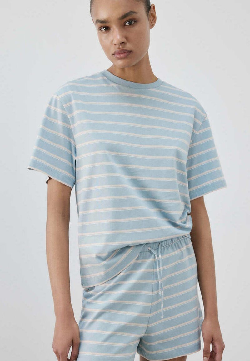 Femme portant une chemise à manches courtes rayée bleu clair et blanc et un short à cordon assorti sur fond uni.