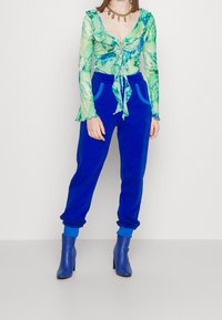 Lightweight, sheer green and blue patterned long-sleeve top with ruffled edges; paired with bright blue joggers and matching ankle boots.