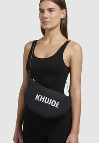 Geantă de umăr din nylon negru, cu o formă rotunjită, având un logo alb "KHUJO 2001" și o curea ajustabilă. Textură netedă, cu hardware minim.