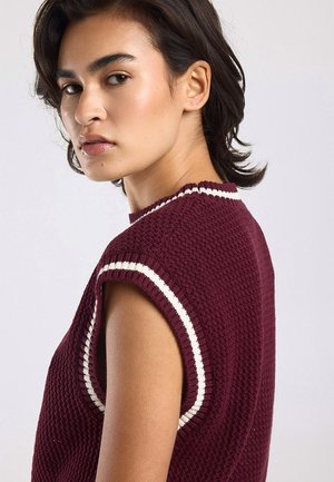 Jong persoon met kort donker haar, die een mouwloos bordeauxrood gebreid topje met witte bies draagt en over de schouder kijkt.