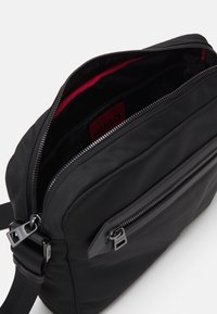 Sort nylon skuldertaske med en lynlåset hovedrum og forlommer, prydet med en rød indvendig foret og metalbeslag.