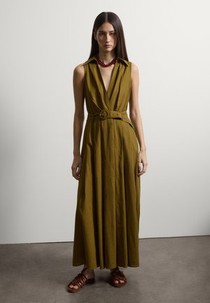 Vrouw staat tegen een neutrale muur, draagt een mouwloze olijfgroene jurk met ceintuur, een rood kralenketting en bruine geweven sandalen.