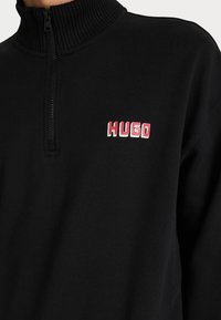 Melna ceturtdaļa-zip sporta krekls ar ribotu apkakli un izšūtu "HUGO" logo rozā un baltā krāsā kreisajā krūtī. Mīksta auduma tekstūra.