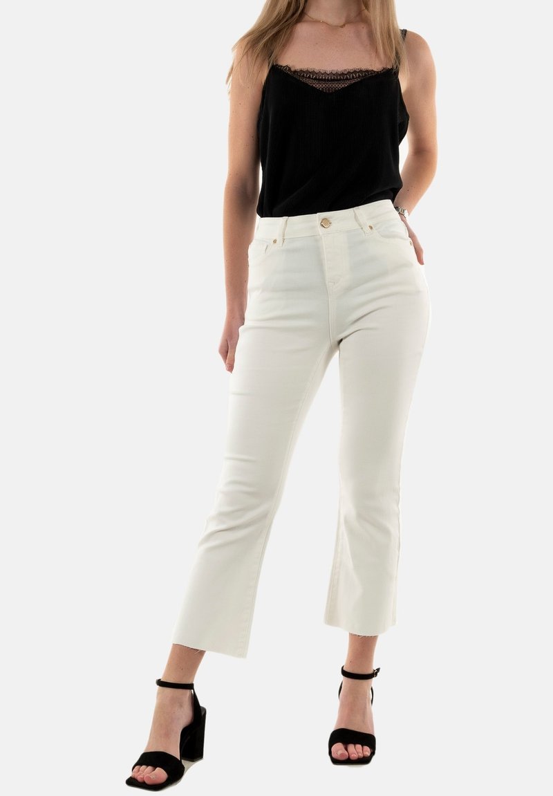 Jeans blancos de talle alto con pernera acampanada, hechos de tela de mezclilla. Llevados con una blusa negra con ribetes de encaje y sandalias de tacón negras.