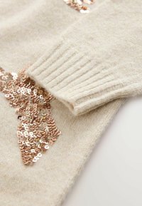Beige-neule, jossa on pehmeä ja teksturoitu pinta. Siinä on suuret ruusukultaiset paljetit ja röyhelömannertuvat, jotka lisäävät visuaalista kiinnostavuutta ja yksityiskohtia.