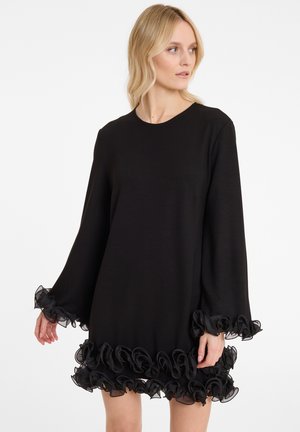 Frau in einem schwarzen langärmeligen Kleid mit Organza-Rüschendetails an den Ärmeln und dem Saum, die auf einem schlichten weißen Hintergrund nach rechts schaut.