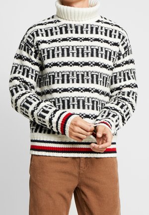 Mężczyzna w czarno-białym swetrze z golfem, wzorzystym, z czerwonymi i granatowymi paskami na mankietach i dolnej części, w połączeniu z brązowymi spodniami sztruksowymi.