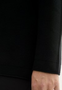 Czarny sweter o strukturze teksturowej z prążkowanym wzorem i gładkim mankietem u dołu. Materiał wydaje się gruby i trwały.