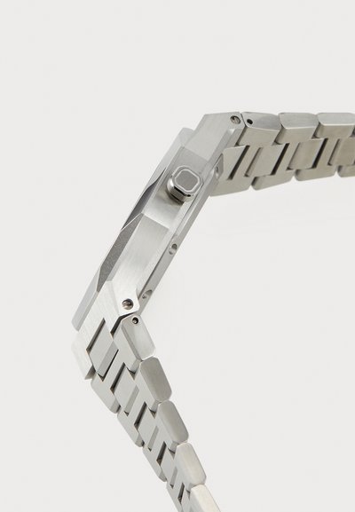 Stainless steel watch with a geometric case design. Silver color with a textured finish and a square push button on the side.