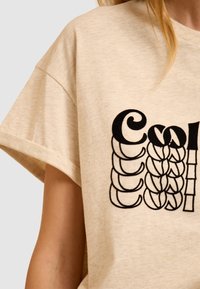 T-shirt oversize beige en tissu doux avec un design graphique noir comprenant du texte stylisé et des formes sur le devant.