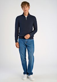 Maritimer, gestrickter Pullover in Marineblau mit einem Viertelreißverschluss-Kragen, kombiniert mit blauen Jeans und weißen Sneakern. Der Pullover hat ein strukturiertes Muster.