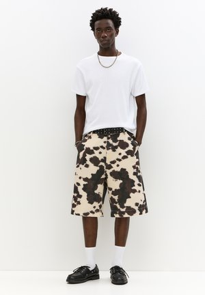 Knelange, køer mønster shorts i beige og sort, parret med en hvid crew neck t-shirt. En sort bælte og sorte sko fuldender outfittet.