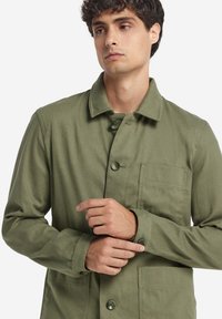Grön, knäppt jacka i texturerat tyg. Har krage, bröstficka och knappdetaljer på ärmarna. Avslappnad, strukturerad design.