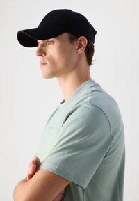 Boné preto e t-shirt de manga curta verde-claro. A t-shirt tem uma textura suave com um pequeno logótipo no peito. Vista de perfil do modelo.