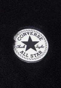 Weißes, rundes Converse All Star Logo-Patch mit einem schwarzen Stern und dem Text "Chuck Taylor" auf einem schwarzen, flauschigen Stoffhintergrund.