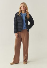 Une femme aux longs cheveux blonds porte une veste matelassée noire avec une doublure à motifs bleus, un pull bleu, un pantalon marron à jambes larges et des chaussures beige.