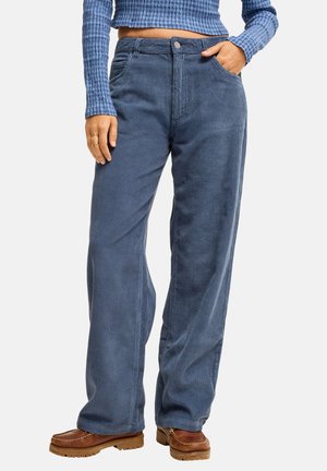 Femme portant un pantalon large en velours côtelé bleu, des chaussures marron et un crop top côtelé à manches longues bleu, avec une main dans la poche.