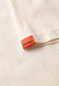 Cremefarvet stof med en fin tekstur, der har en orange vævet etiket med "SUPERDRY" broderet i sort.