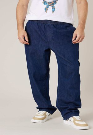 Mann trägt lockere dunkelblaue Jeans mit elastischem Bund und weißen Turnschuhen, steht vor einem einfarbigen Hintergrund.