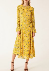 Femme portant une robe midi jaune à manches longues et imprimé floral, associée à des sandales noires à talons hauts et bouts ouverts.