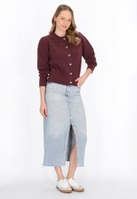 Burgundijas adīta jaka ar sirds formas pogām, apvienota ar gaiši zilu denim svārku ar priekšējo šķēlumu un saplēstām malām, nēsāta ar baltām sporta apaviem.