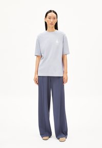 Lichtgrijze oversized t-shirt met korte mouwen en een klein wit logo; gecombineerd met wijd uitlopende marineblauwe broeken en beige schoenen.