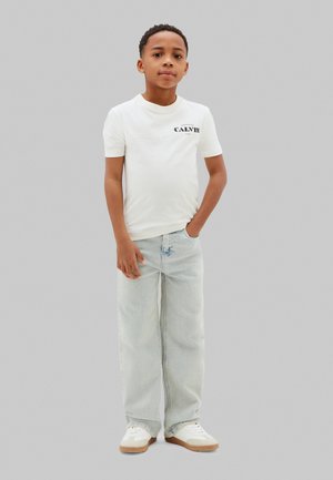 Dreng stående med den ene hånd i lommen iført en hvid Calvin Klein t-shirt, lyseblå vide jeans og hvide sneakers på grå baggrund.