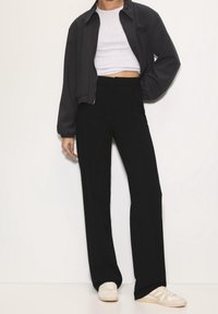 Personne portant un pantalon droit noir taille haute, un t-shirt blanc court, une veste noire zippée et des baskets blanches, debout devant un fond uni.