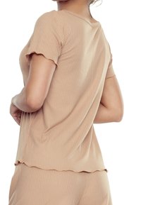 Modelka w jasnobrązowej, prążkowanej bluzce z krótkim rękawem, przyjmuje zrelaksowaną pozę. Tył bluzki ma koronkowy brzeg.