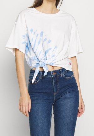 Valkoinen puuvillapaita, jossa on sininen sidontakuosi ja solmittu etuosa, yhdistettynä tummansinisiin farkkuihin. Lyhyet hihat, rento malli.