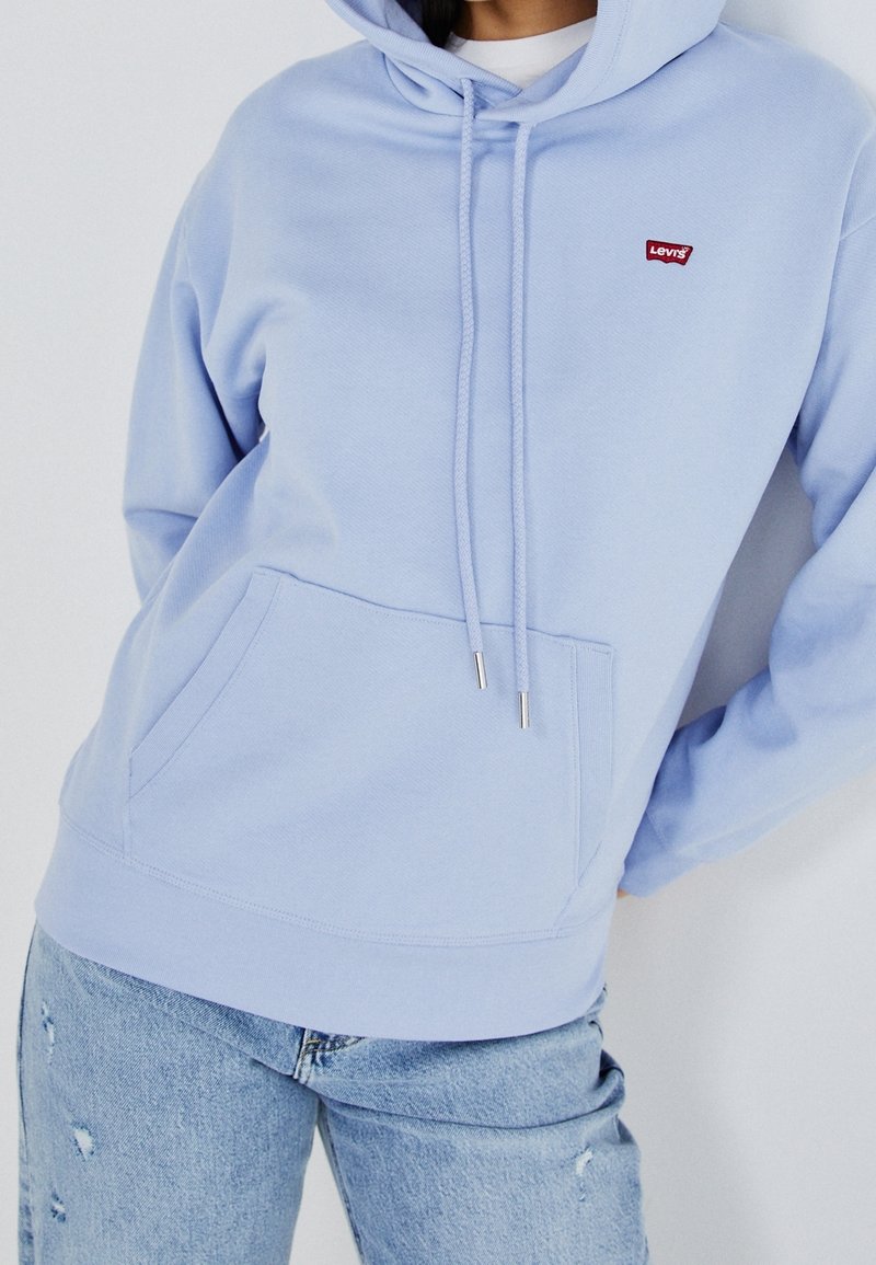 Sudadera de color azul claro con bolsillo frontal, capucha con cordón y logo rojo. La tela parece suave; cuenta con mangas acampanadas y un corte relajado.