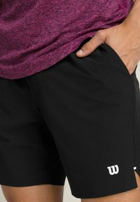 Schwarze Sportshorts mit glatter Textur, ausgestattet mit einem weißen Logo auf der linken Seite und einer Seitentasche, kombiniert mit einem lila strukturierten Oberteil.