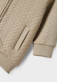 Giacca trapuntata beige con motivo a spina di pesce, dotata di polsini a coste e tasca laterale, realizzata in morbido tessuto a maglia.