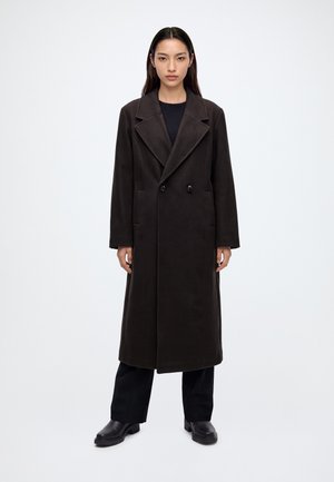 Femme debout face à nous, portant un long manteau sombre à double boutonnage, un pantalon noir et des chaussures noires sur un fond blanc uni.