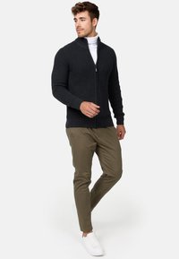 Mørkegrå strikket zip-up genser med ståkrage; kombinert med olivenfargede chinos og hvite sneakers. Modellen poserer avslappet.