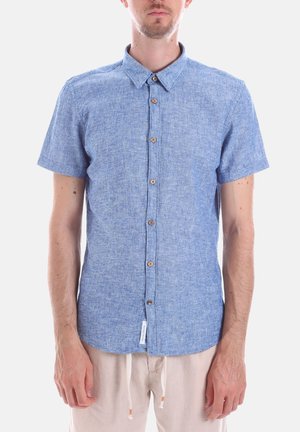 Uomo che indossa una camicia blu a maniche corte con bottoni in legno e pantaloni beige chiaro con coulisse, su sfondo bianco.