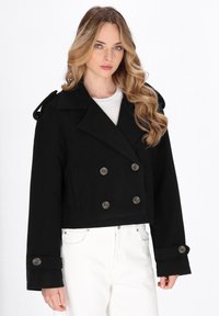 Musta lyhyt peacoat, jossa on tuplaknopit, leveät kaulukset ja olkavuoret. Materiaali näyttää pehmeältä ja sileältä.