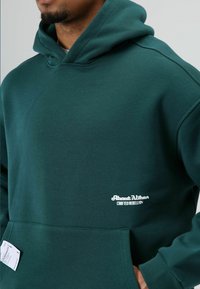 Groen sweatvest met capuchon gemaakt van zachte stof, met een voorzak en een wit "About Vibes" logo op de borst.