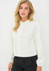 Hvid skjorte med knapper og lodrette plisseringer foran og på ærmerne. Har en lige krave og let puffede lange ærmer.