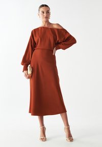 Midi šaty v rustikálnej farbe vyrobené z hladkej látky, s dizajnom bez ramienok a habrovými rukávmi, doplnené textúrovanou zlatou kabelkou.