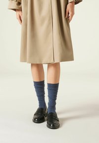 Schwarze Slipper mit strukturierter Oberfläche, kombiniert mit gerollten blauen Socken, getragen unter einem beigen Trenchcoat. Der Mantel hat ein klares, gerades Design.