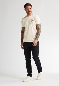 Camiseta de algodón beige con estampado gráfico en el pecho, combinada con pantalones cargo negros y zapatillas blancas, de pie contra un fondo liso.